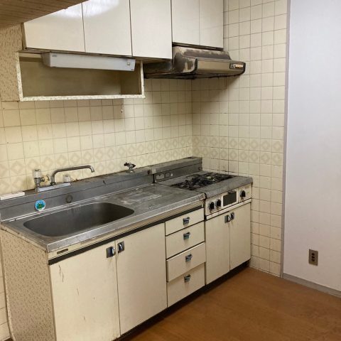 キッチンリフォーム前の状態。経年劣化したブロックキッチンで、換気扇やガスコンロ周辺に油汚れやサビが見られ、壁のタイルや収納棚も古さを感じさせる。